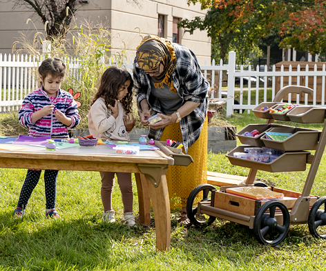 Two children doing a craft on an outdoor table instructed by a teacher